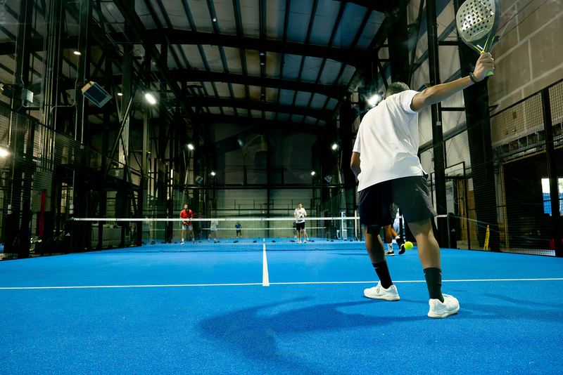 A player serves under the steel trusses of Foundry Padel's warehouse courts